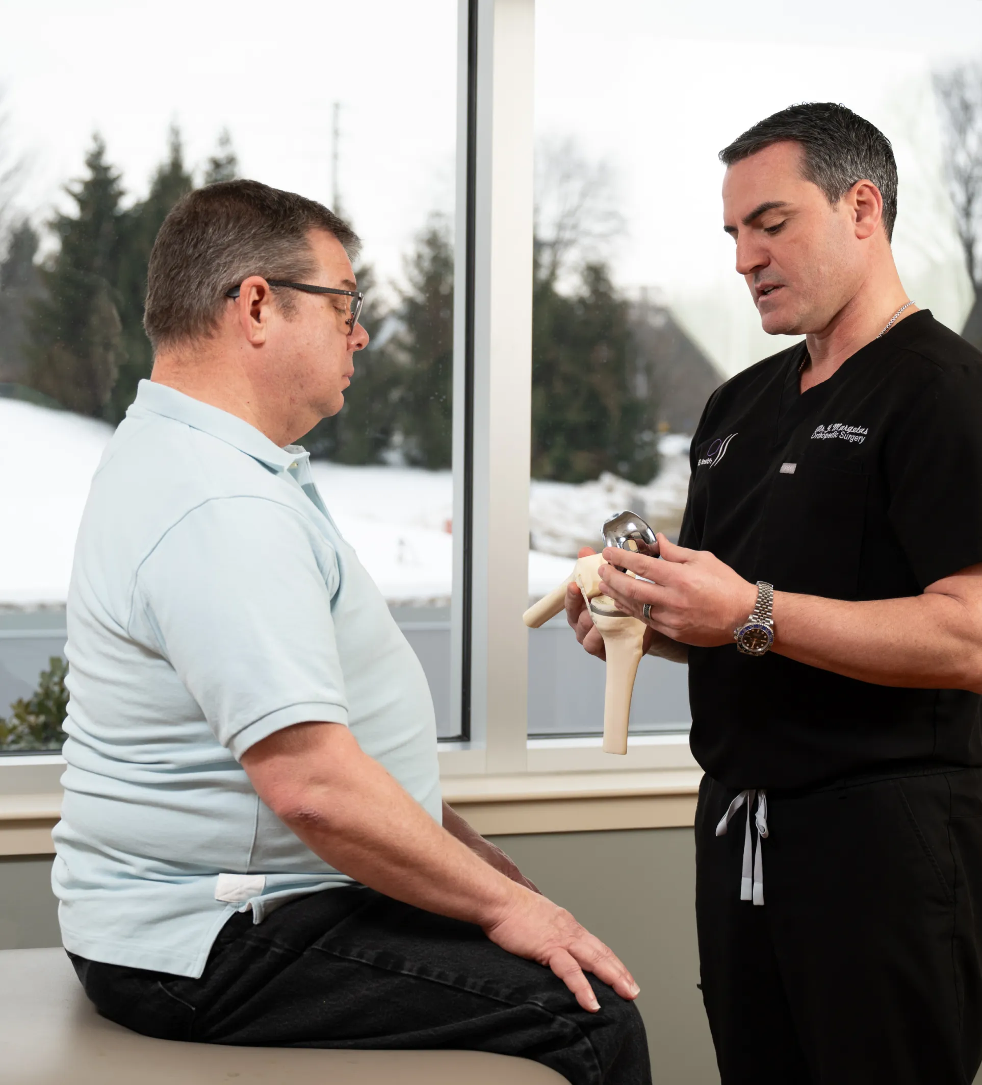 An OSS Health surgeon, holding a knee replacement model, gives a patient a knee replacement consultation.
